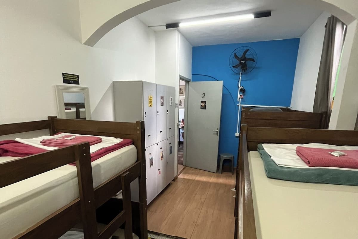 A dormitory room with bunk beds, a desk, a chair, a mirror, a wardrobe, and a blue wall with a fan.