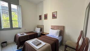A hotel room with two single beds, a small table, a chair, and a window with a view of buildings and trees. - Rota BH Hostel (Belo Horizonte)