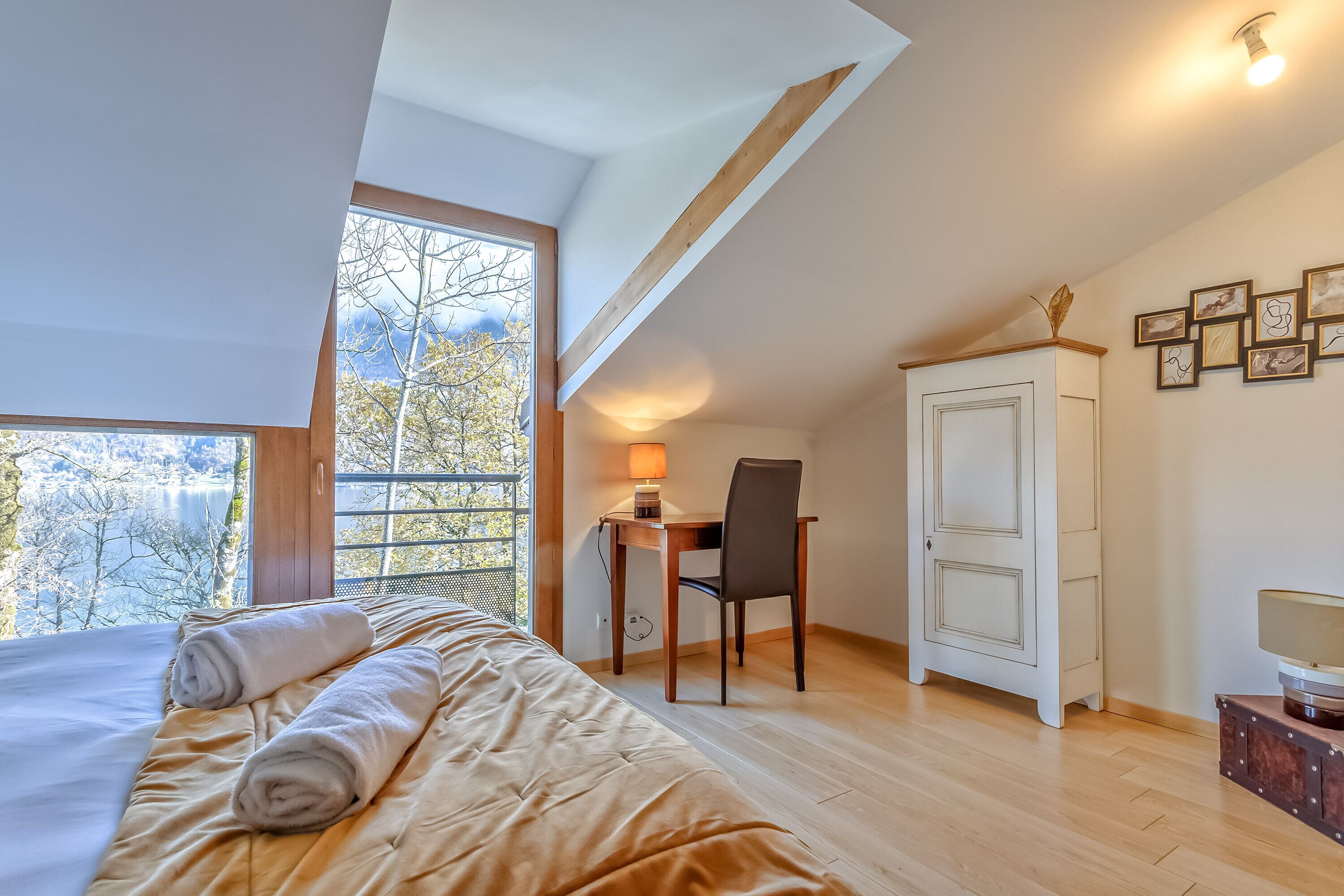 A bedroom with a large bed, a desk with a chair, a wardrobe, and a window with a view of trees and a lake.