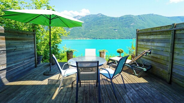 A wooden deck with a table and chairs, a green umbrella, and a view of a lake and mountains. - Les Terrasses de Bredanne (Doussard)