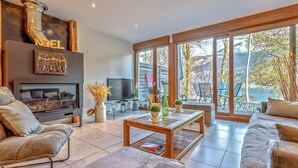 A modern living room with a fireplace, comfortable seating, a coffee table, and large windows offering a view of the outdoors. - Les Terrasses de Bredanne (Doussard)