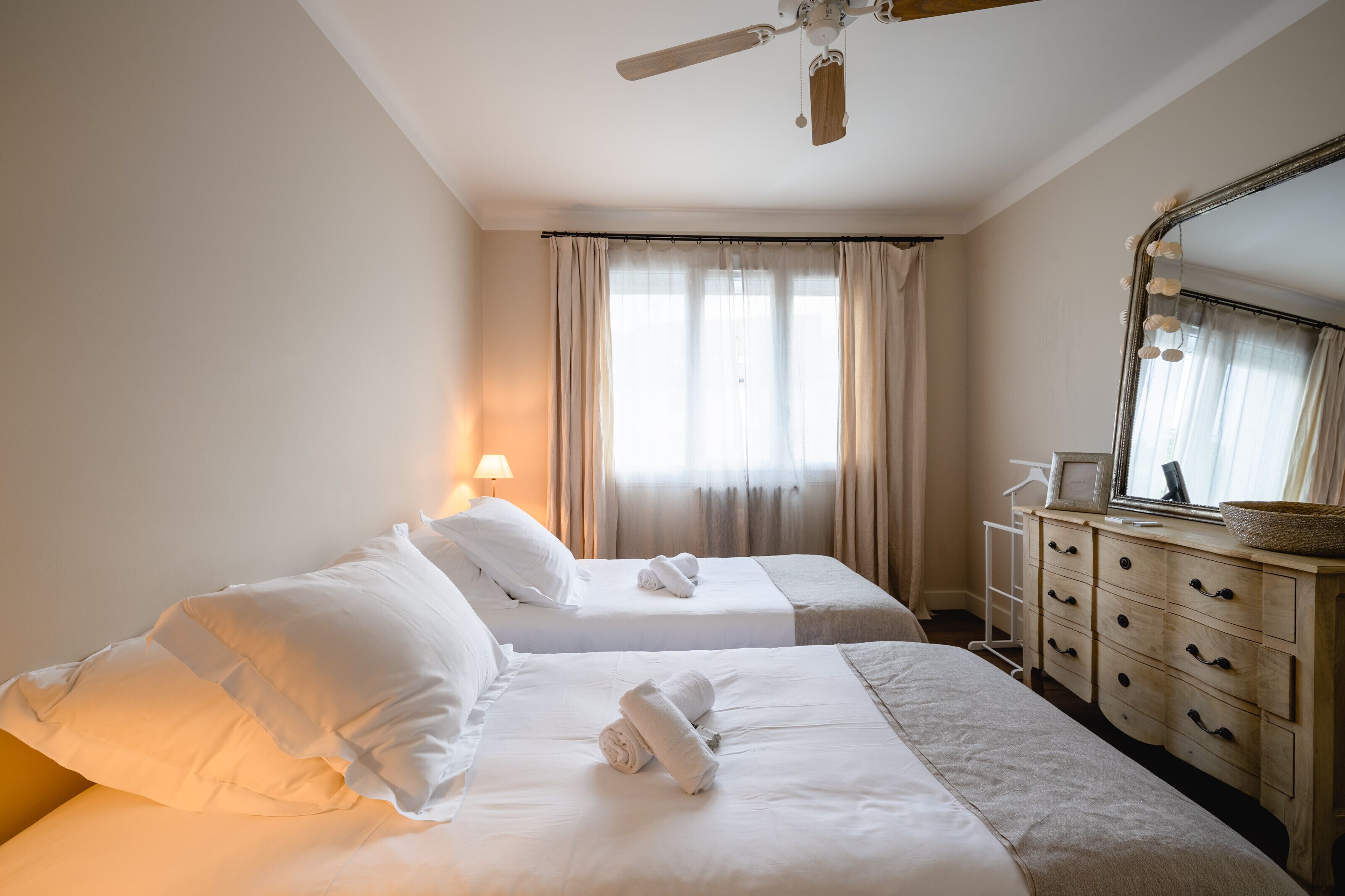 A hotel room with two beds, a dresser, a ceiling fan, and a large mirror with lights.