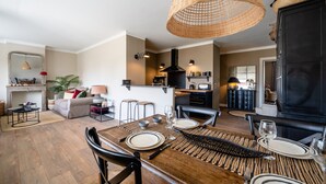 A dining area with a wooden table set for four, surrounded by chairs. A kitchen area is visible in the background with a black stove and various kitchen items. - LA MAVÉRIA – Elegant 100 m² flat near Lake Annecy (Annecy-le-Vieux)