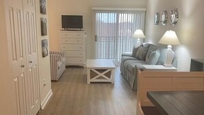Amplia sala de estar con sofá, mesa de centro y televisor sobre un soporte. Hay una puerta corrediza de vidrio que da a un balcón.