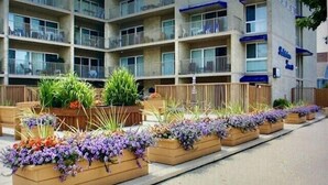 Una hilera de macetas de madera con diversas flores y plantas a lo largo de una acera frente a un edificio de varios pisos con balcones.