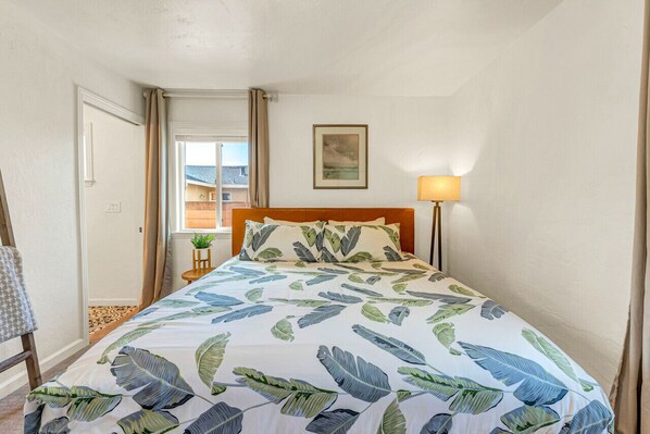 Une chambre à coucher avec un lit, une table de chevet, une lampe, une fenêtre avec des rideaux et un tableau au mur.