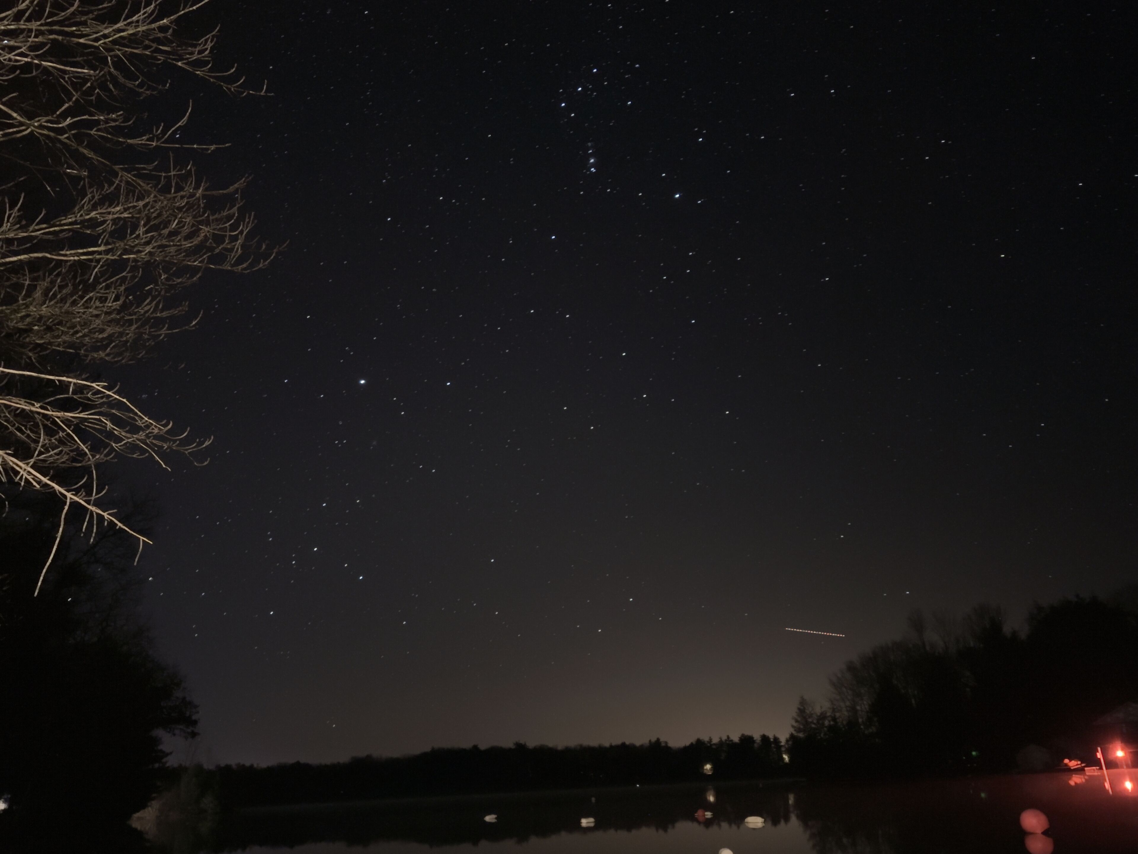 Stjerneklar nattehimmel over stille vand, omgivet af træer og en svagt synlig vej med lys.