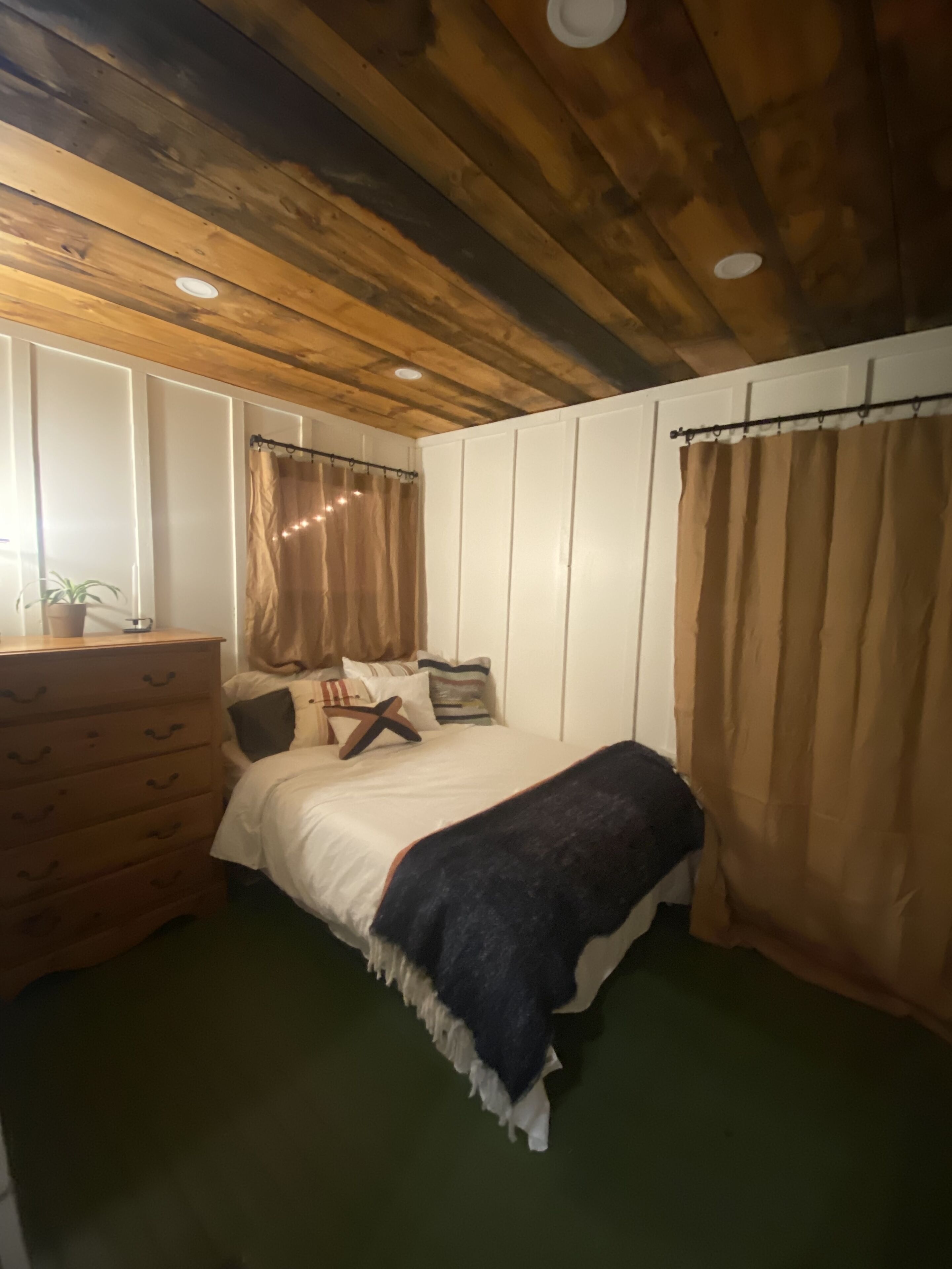 A bedroom with a wooden ceiling, a bed with a white duvet, a wooden dresser, and a window with curtains.