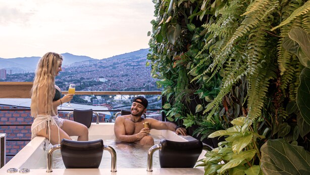 Un jacuzzi en la azotea con dos personas, rodeado de vegetación exuberante y vistas panorámicas de la ciudad.