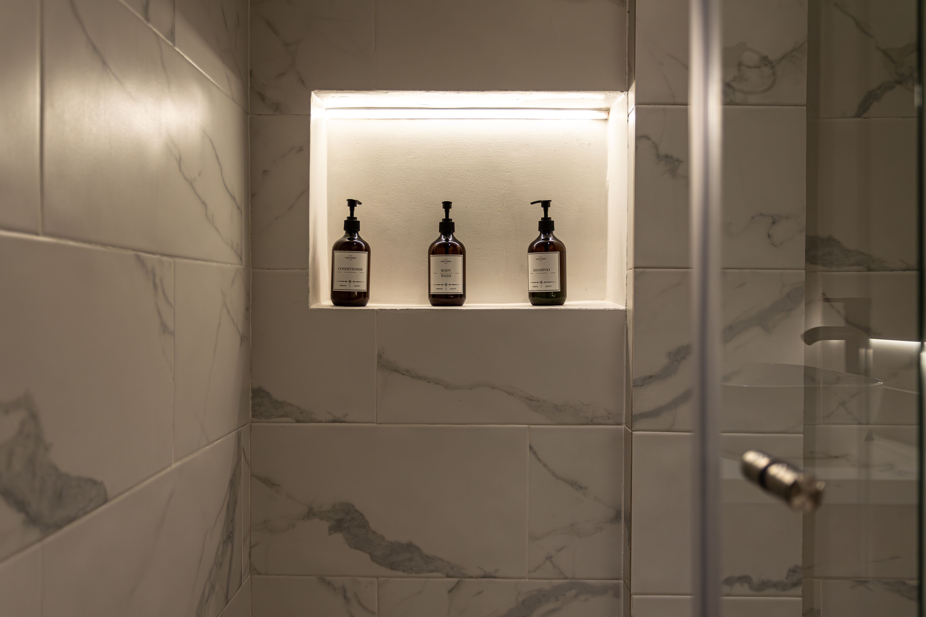 A bathroom with marble tiles, a shelf with three bottles of shampoo, and a glass shower enclosure.