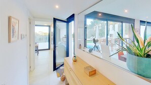 Um interior moderno com um espelho grande refletindo uma piscina e mobiliário de exterior, um banco de madeira com objetos decorativos, e uma porta que dá para outro quarto.