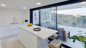Uma cozinha moderna com uma ilha central, sala de jantar com mesa e cadeiras, e grandes janelas com vista para o verde.