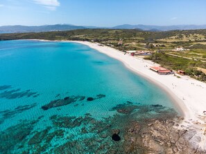 Ein Luftbild einer Küstenregion mit Strand, klarem türkisblauem Wasser, felsigen Abschnitten und vereinzelten Gebäuden.