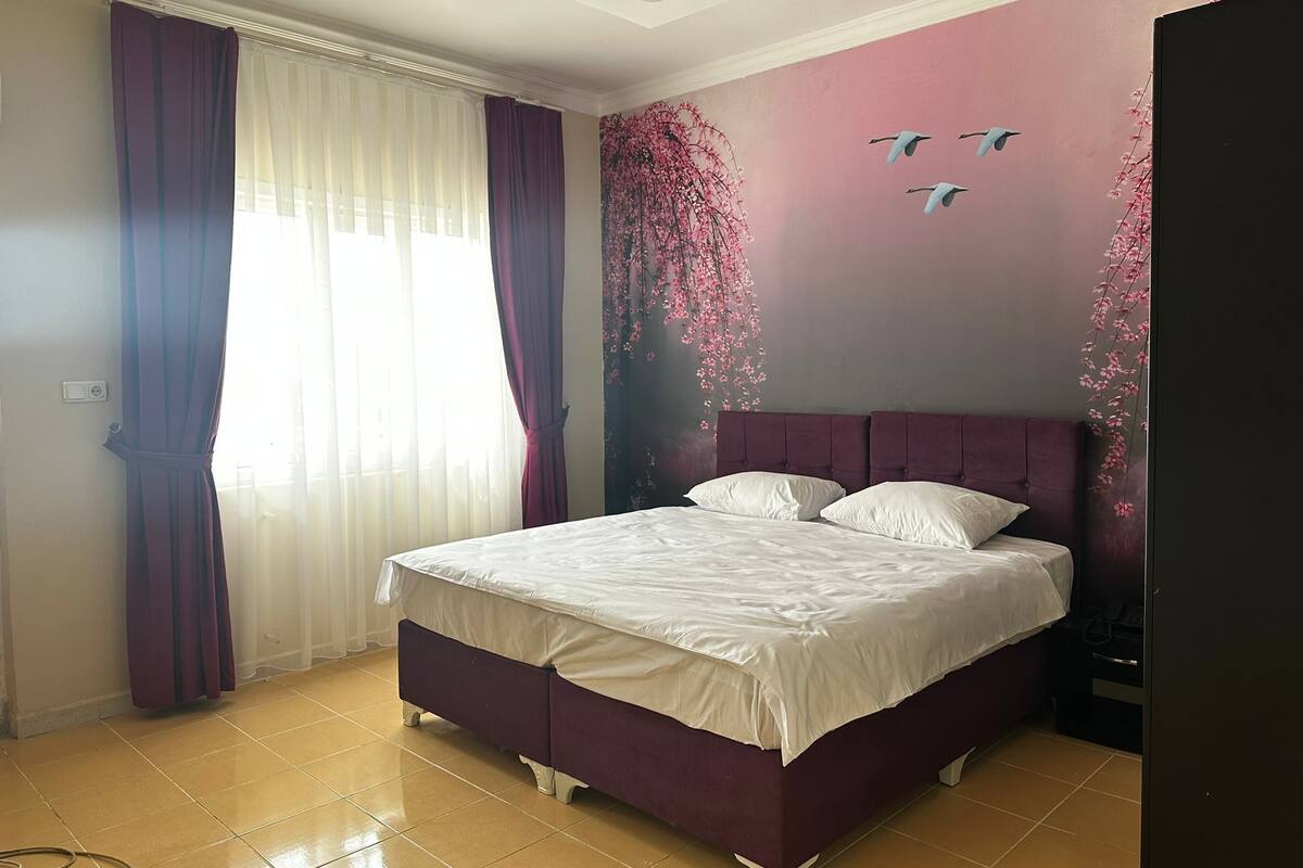 A bedroom with a bed, curtains, a wall mural, and a tiled floor.