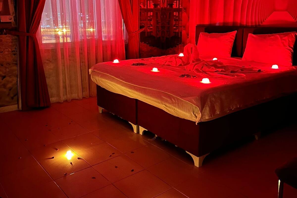 A dimly lit bedroom with a large bed, red curtains, and a view of the cityscape through the window.