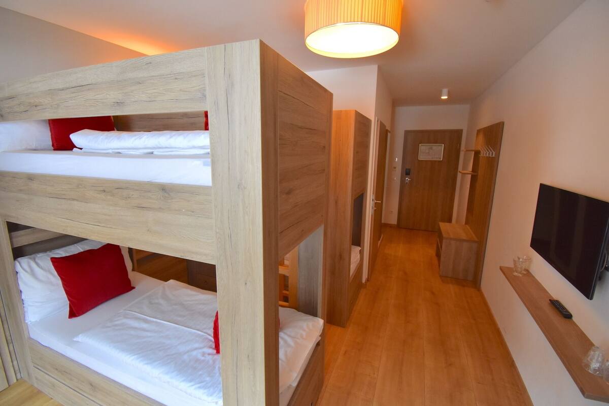 A room with bunk beds, a wooden floor, a hallway, a door, and a TV mounted on the wall.