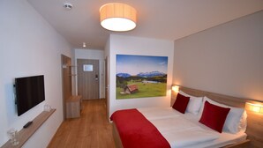 Habitación de hotel con cama, televisor, un cuadro de un paisaje y un pasillo que conduce a una puerta.