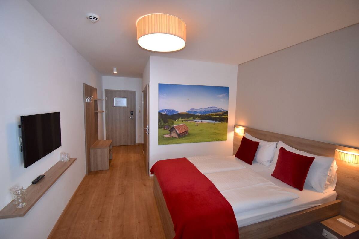 A hotel room with a bed, a TV, a painting of a landscape, and a hallway leading to a door.