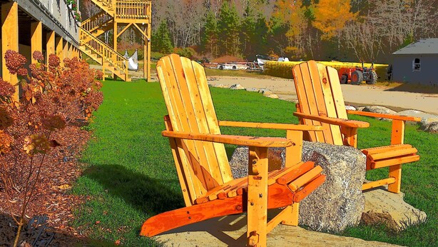 Zwei hölzerne Adirondack-Stühle mit Steinuntersetzung, ein Holzboden und eine gelbe Treppe, die zu einem Gebäude führt.
