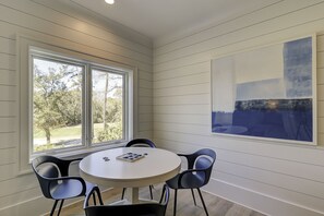 A dining area with a round table and four chairs, large windows with a view of trees, and a large abstract painting on the wall. - 51 Mooring Buoy-New Home w/elevator & AMAZING POOL, 2 min walk to the Beach (Hilton Head Island)