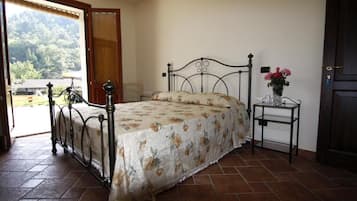 Un dormitorio con estructura de cama metálica, colcha floral, mesita de noche con un florero y vistas al exterior a través de una puerta abierta.