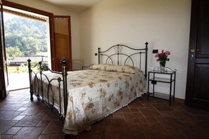 Ein Schlafzimmer mit einem Metallbettgestell, einer blumigen Bettdecke, einem kleinen Nachttisch mit Blumen und Blick ins Freie durch eine offene Tür.