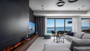 Un salon moderne doté d’une cheminée, d’un grand téléviseur à écran plat et d’un canapé placé face à une grande fenêtre offrant une vue sur la mer.