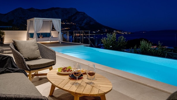 Un espace aménagé au bord de la piscine, avec un canapé en osier, une table ronde en bois sur laquelle sont posés une bouteille de vin et un verre, et une vue sur les montagnes la nuit.