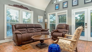 A living room with a brown leather sofa, a patterned armchair, a glass coffee table, and a blue vase. - The Island Spot - 721 Bienville 3 Bedroom Home by RedAwning (Dauphin Island)