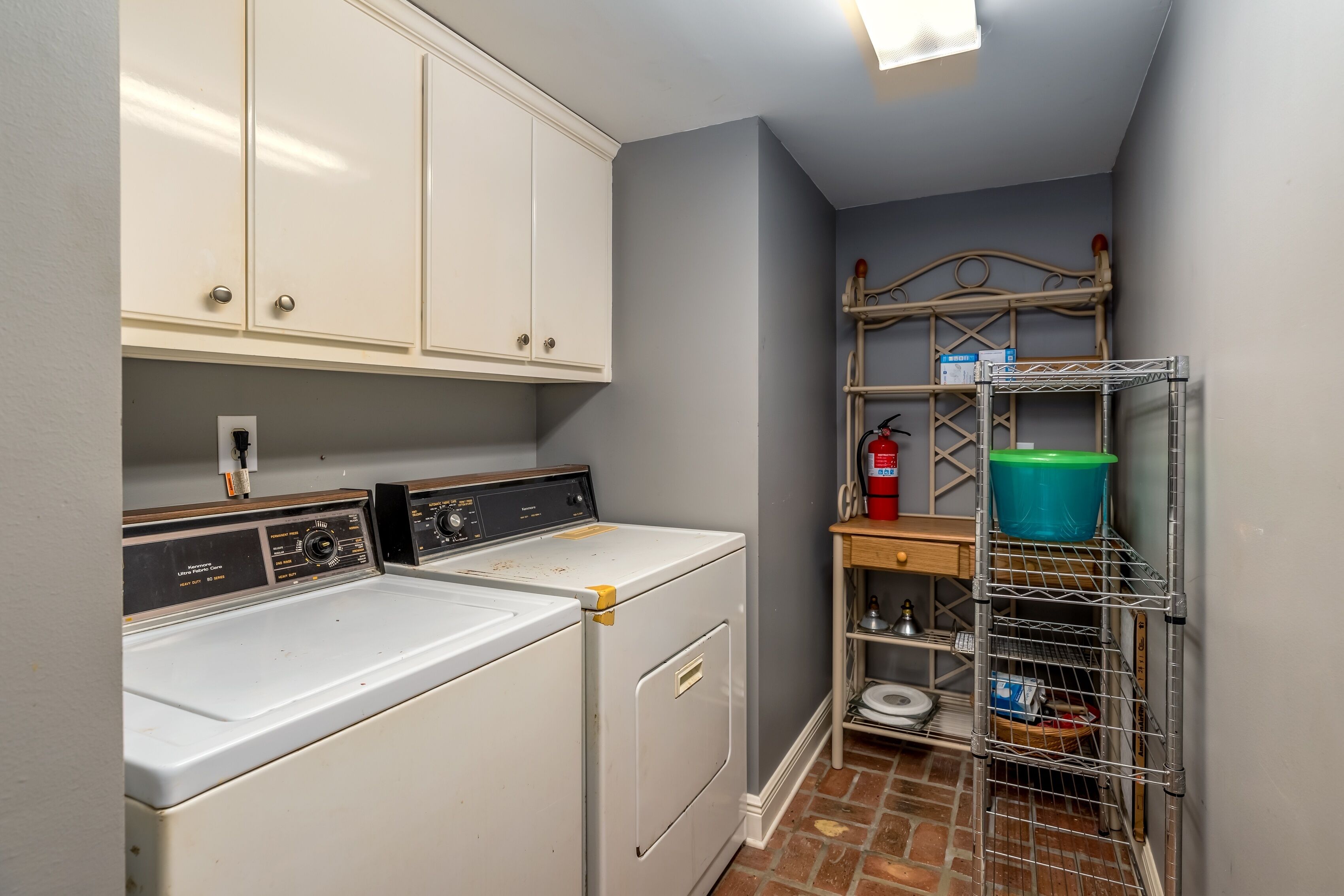 A laundry area with a washing machine, dryer, and metal shelving unit with a blue bucket.