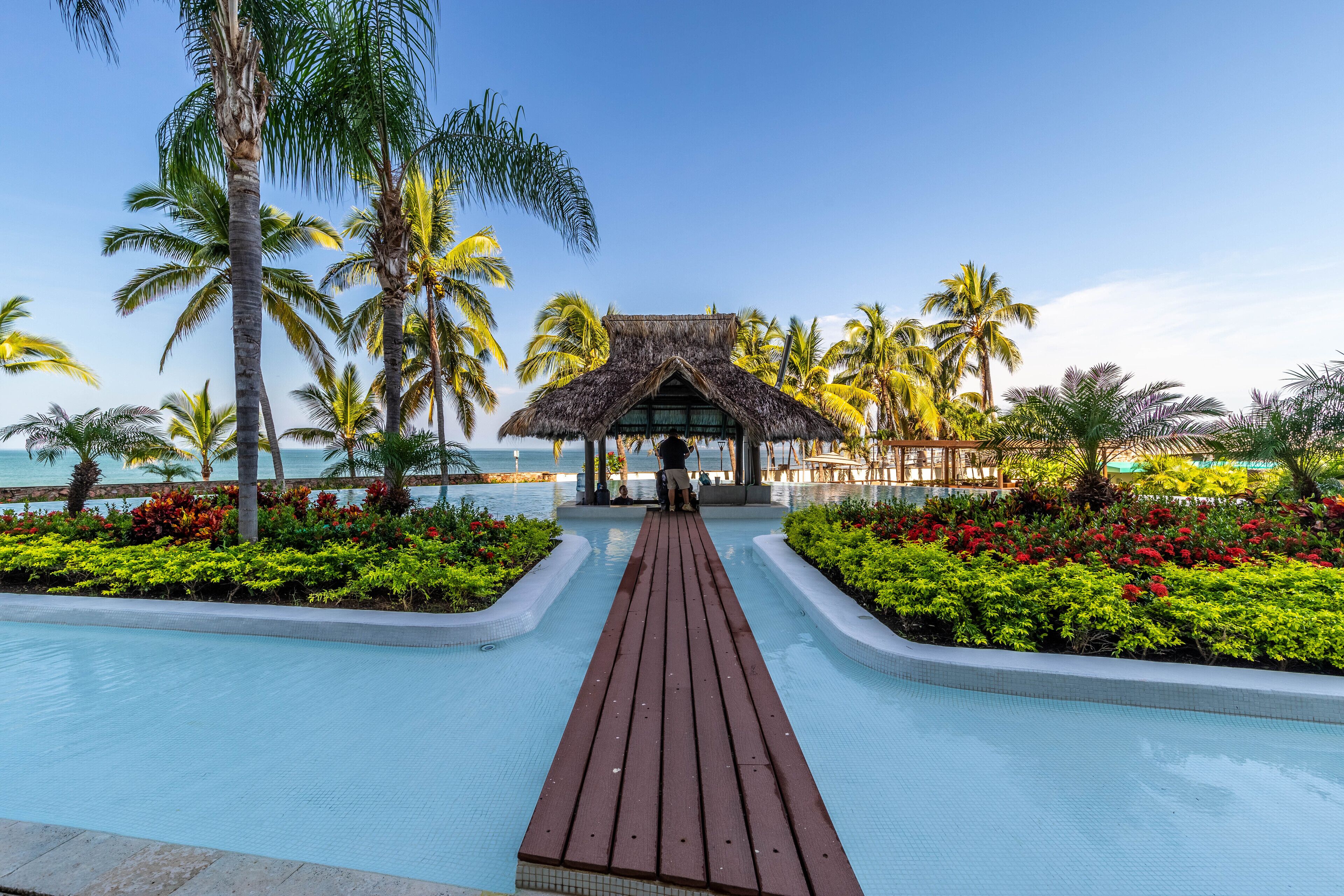 A wooden pathway leading to a thatched-roof structure surrounded by palm trees and a pool.