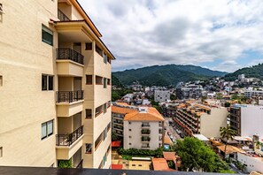 Un immeuble à plusieurs étages doté de balcons, offrant une vue sur un paysage urbain avec des montagnes en arrière-plan, et un ciel dégagé.