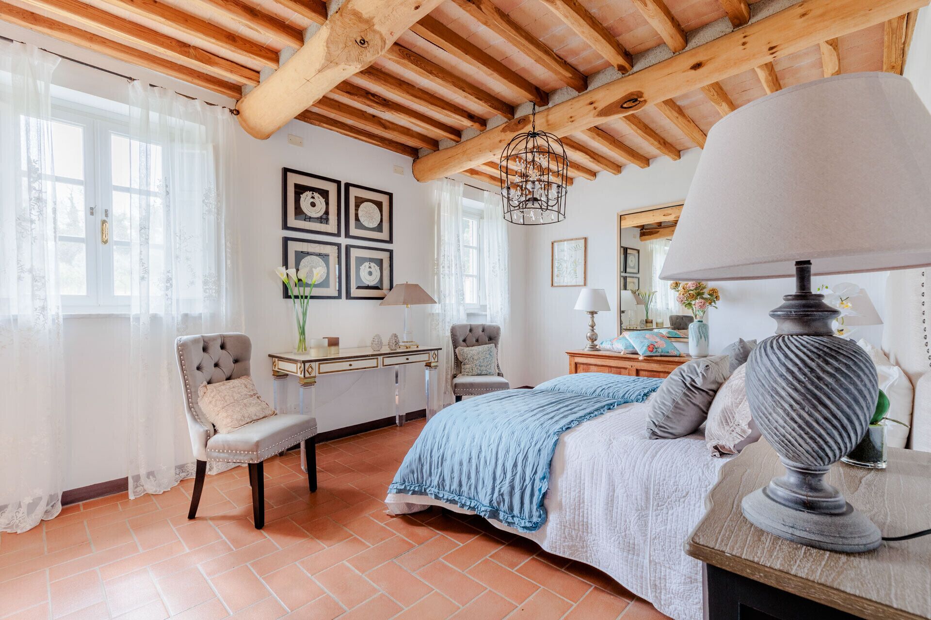 A bedroom with a wooden ceiling, a bed with a blue blanket, a chair, a desk, and a chandelier.