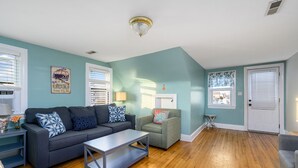 A living room with a grey sofa, a small table, and a green armchair. There are two windows with blinds, a door, and a ceiling light fixture.