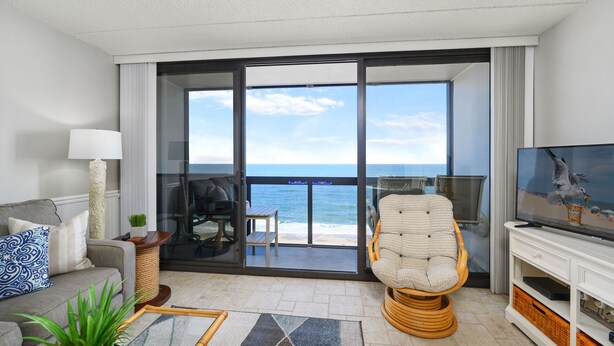 A modern living room with a view of the ocean, a comfortable sofa, a wicker chair, a television, and a glass door leading to a balcony.