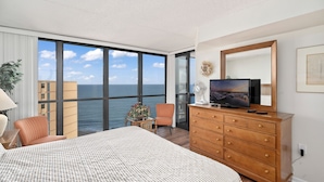 A hotel room with a large bed, a dresser, a television, and a view of the ocean through large windows. - Golden Sands 1901 by Seaside Vacations & Sales (Ocean City)