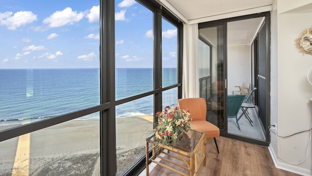 A balcony with a view of the ocean, a wicker chair, and a table with a floral arrangement. - Golden Sands 1901 by Seaside Vacations & Sales (Ocean City)