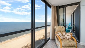 A balcony with a view of the beach, a wicker chair with cushions, and a glass door leading to a balcony area with blue chairs. - Fabulous Oceanfront condo w/ pools by Seaside Vacations & Sales (Ocean City)