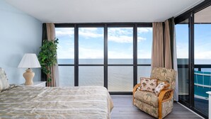 A hotel room with a large bed, a chair, a lamp, and a view of the ocean through large windows. - Fabulous Oceanfront condo w/ pools by Seaside Vacations & Sales (Ocean City)