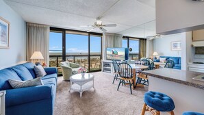 A hotel room with a large window, a ceiling fan, a sofa, a dining table, and a kitchen area.
