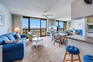 A hotel room with a large window, a ceiling fan, a sofa, a dining table, and a kitchen area.