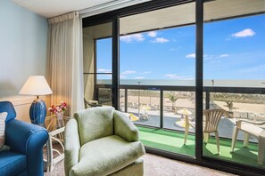 A room with a large window, a blue chair, a green armchair, a lamp, and a small table with flowers.