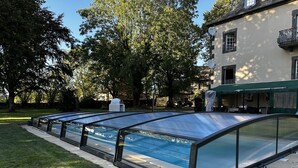 A swimming pool with a glass enclosure, surrounded by grass and trees, with a building in the background.