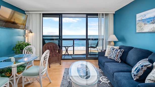 A modern living room with a view of the ocean, a sofa, a coffee table, and a dining area with chairs and a glass table.