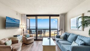 A living room with a sofa, armchair, and coffee table, a TV, and a view of the beach through large windows.