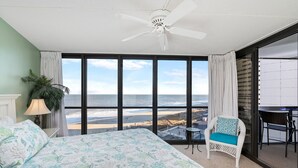 A bedroom with a large bed, a chair, a table, and a view of the beach.