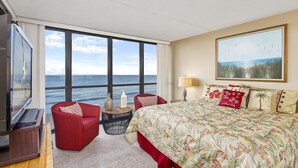 A hotel room with a large window offering a view of the ocean, a bed with floral bedding, a red armchair, a small side table, and a television. - Golden Sands 912 by Seaside Vacations & Sales (Ocean City)