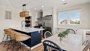 Una cocina moderna con una isla central, armarios blancos, electrodomésticos de acero inoxidable y vistas a casas por la ventana.