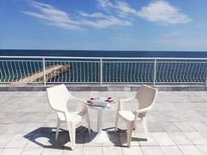 A terrace with two white plastic chairs and a small table, overlooking a rocky coastline and the sea.