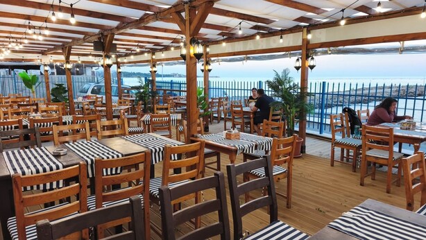 Ein Restaurant im Freien mit Holztischen und -stühlen, Blick auf das Meer und einer hölzernen Pergola.